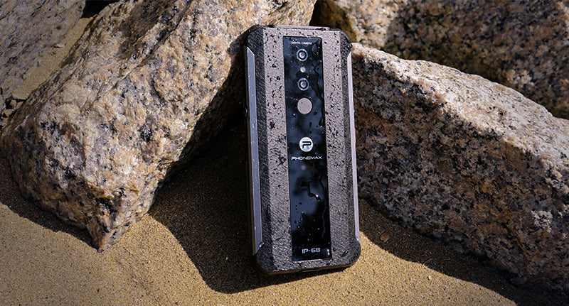 Black Phonemax rugged phone resting between rocks on sandy outdoor terrain