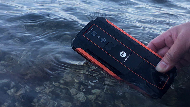 Black and red Phonemax rugged smartphone held partly over clear outdoor water